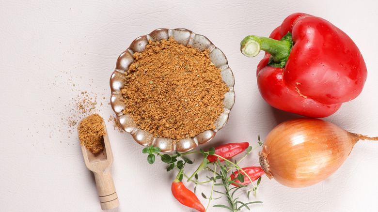 A pot of cajun seasoning, a red pepper, yellow onion, and red chilis.